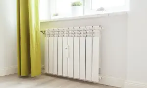 Biały kaloryfer z termostatem przy oknie, obok zielonej zasłony.