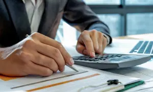 Osoba w garniturze liczy na kalkulatorze, analizując dokumenty. Planuje, kiedy zapłacić podatek za wynajem mieszkania.