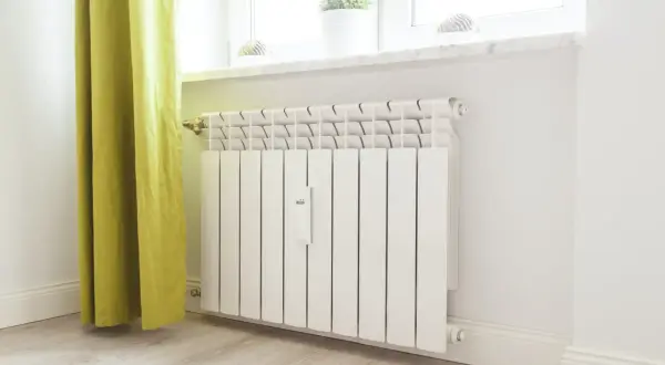 Biały kaloryfer z termostatem przy oknie, obok zielonej zasłony.